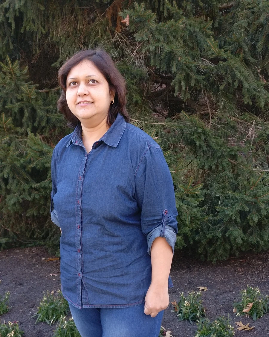 Dr. Komal Aggarwal in a denim shirt, standing in front of evergreen trees.