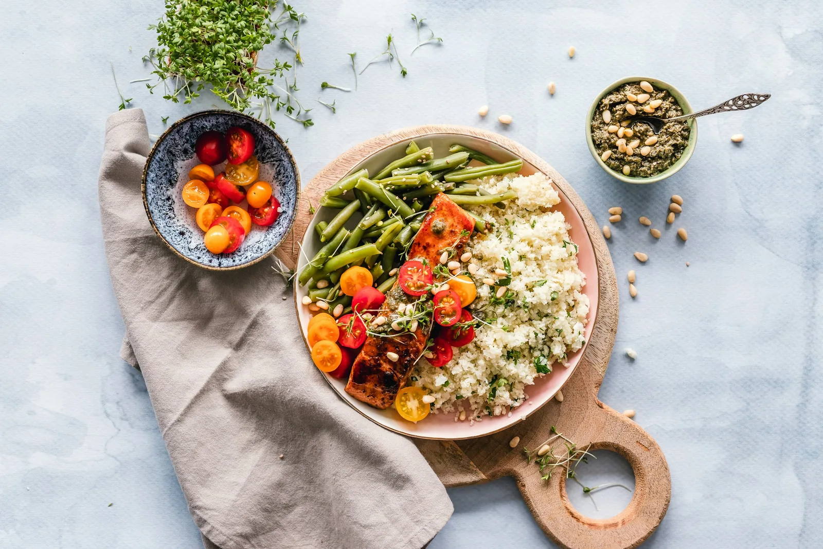 A plate of grains, green beans, roasted tomatoes and peppers on a wooden board, with a bowl of cherry tomatoes and a dish of seeds and sprouts on a pale surface with a linen cloth.