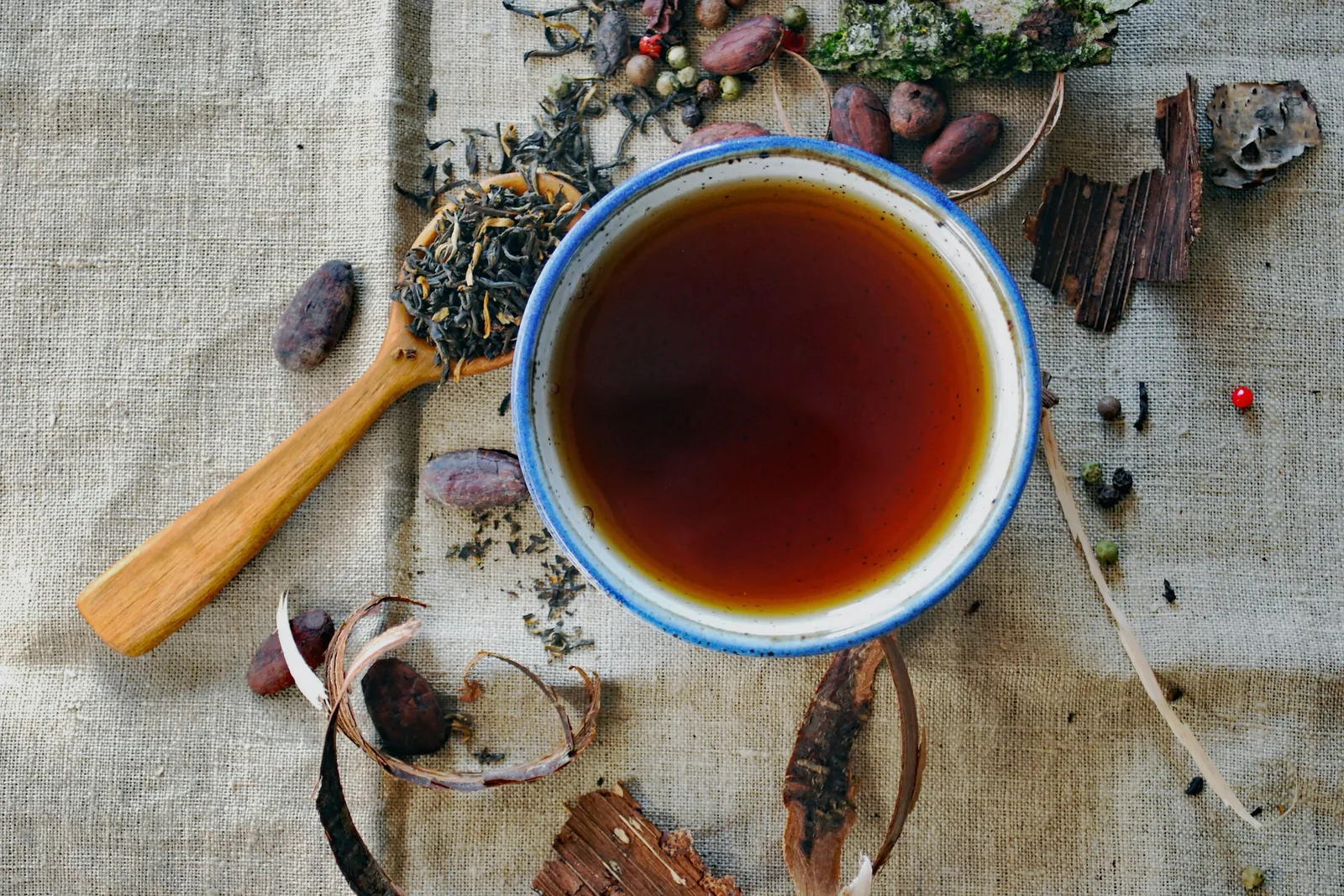 A bowl of dark spiced tea seen from above, surrounded by whole spices, herbs and a wooden spoon on linen.