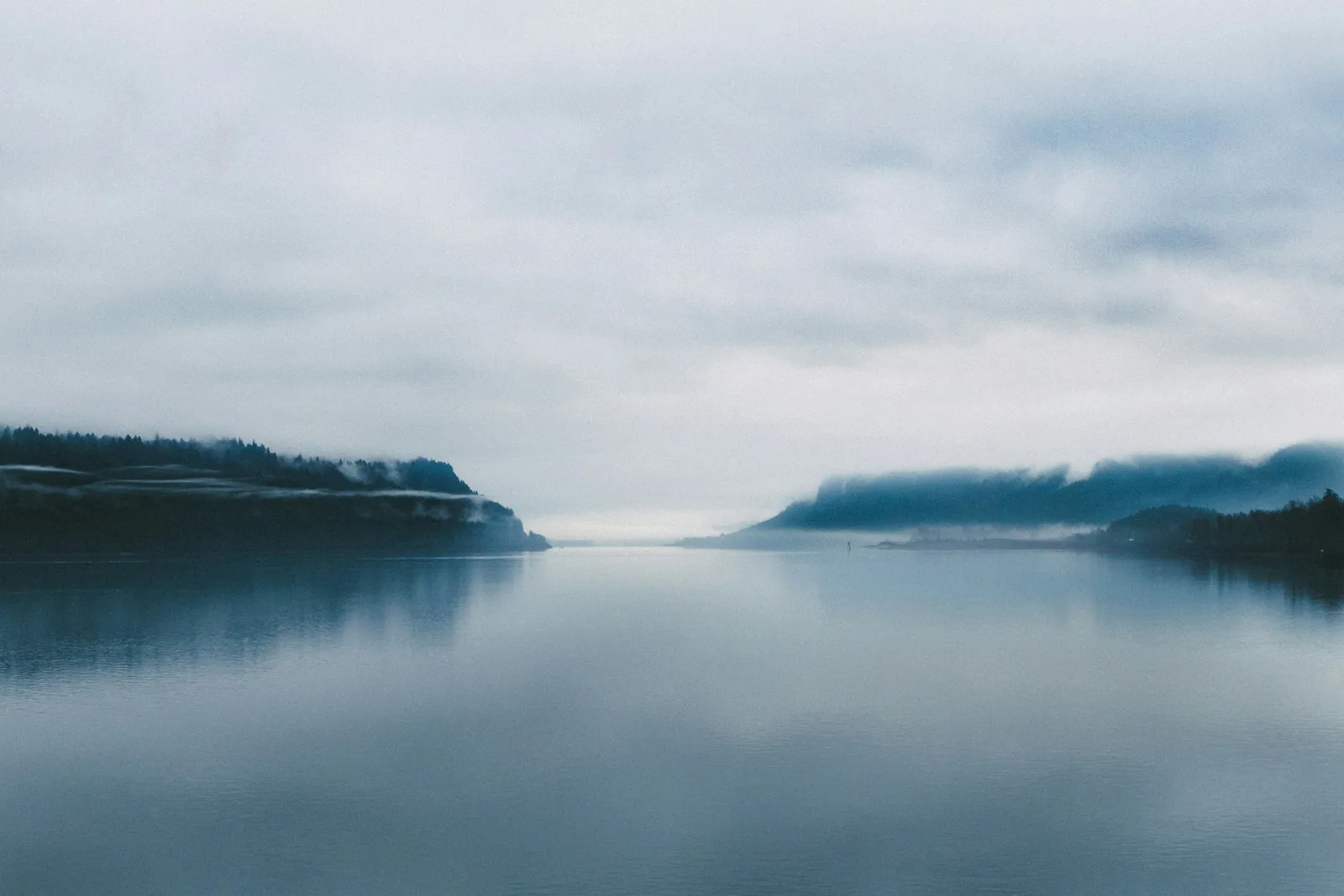 Calm, misty water reflecting a soft grey sky, with forested hills in the distance.